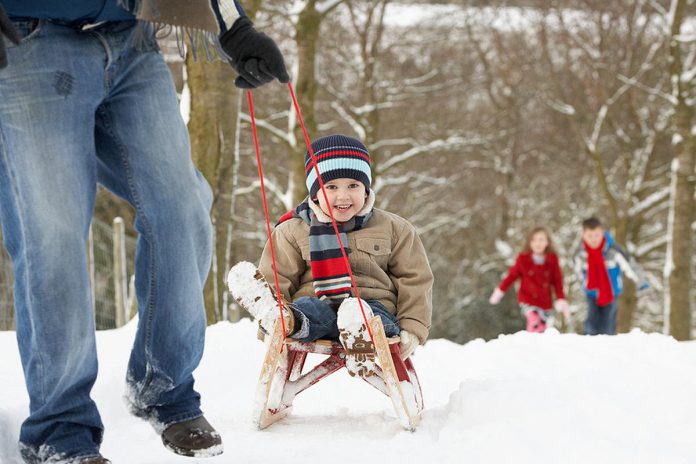 A outdoors winter scene showing a man pulling a young boy on a sled through the woods with a young girl and boy following in the background. (Stock photo)