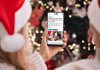 A person wearing a Santa hat in front of a Christmas tree holding a smartphone displaying the 2024 holiday hours listing on kawarthaNOW's website. (Photo: kawarthaNOW)