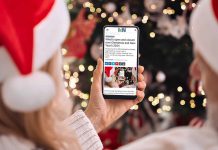 A person wearing a Santa hat in front of a Christmas tree holding a smartphone displaying the 2024 holiday hours listing on kawarthaNOW's website. (Photo: kawarthaNOW)