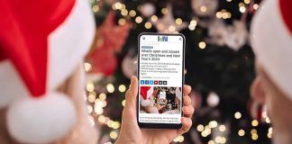 A person wearing a Santa hat in front of a Christmas tree holding a smartphone displaying the 2024 holiday hours listing on kawarthaNOW's website. (Photo: kawarthaNOW)