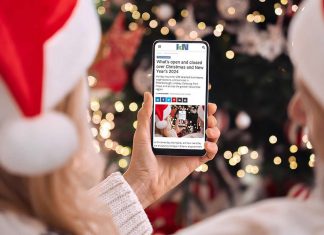 A person wearing a Santa hat in front of a Christmas tree holding a smartphone displaying the 2024 holiday hours listing on kawarthaNOW's website. (Photo: kawarthaNOW)