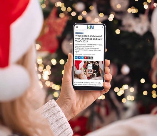 A person wearing a Santa hat in front of a Christmas tree holding a smartphone displaying the 2024 holiday hours listing on kawarthaNOW's website. (Photo: kawarthaNOW)