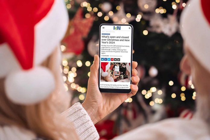 A person wearing a Santa hat in front of a Christmas tree holding a smartphone displaying the 2024 holiday hours listing on kawarthaNOW's website. (Photo: kawarthaNOW)