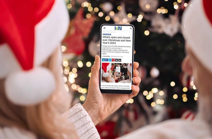 A person wearing a Santa hat in front of a Christmas tree holding a smartphone displaying the 2024 holiday hours listing on kawarthaNOW's website. (Photo: kawarthaNOW)