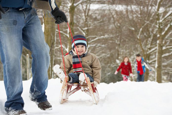 A winter scene showing a man pulling a young boy on a sled with a young girl and boy walking in the background. (Stock photo)