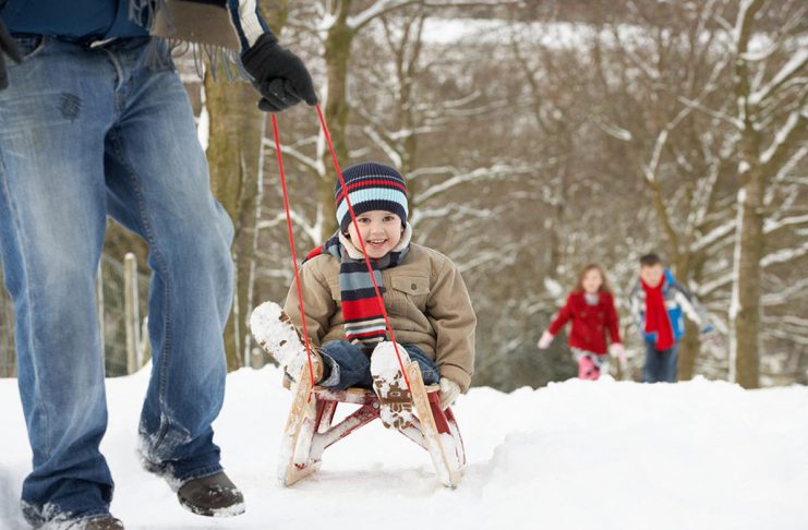 A winter scene showing a man pulling a young boy on a sled with a young girl and boy walking in the background. (Stock photo)