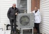 Rick and Annie Lochhead of Norwood stand next to the outdoor unit of their air source heat pump, which they installed in 2023. After doing some other upgrades, they estimate they're saving $50/month on their energy bills. (Photo: Clara Blakelock / GreenUP)