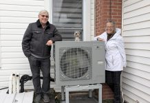 Rick and Annie Lochhead of Norwood stand next to the outdoor unit of their air source heat pump, which they installed in 2023. After doing some other upgrades, they estimate they're saving $50/month on their energy bills. (Photo: Clara Blakelock / GreenUP)