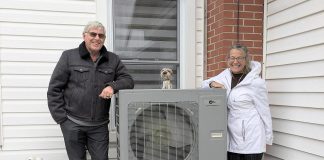 Rick and Annie Lochhead of Norwood stand next to the outdoor unit of their air source heat pump, which they installed in 2023. After doing some other upgrades, they estimate they're saving $50/month on their energy bills. (Photo: Clara Blakelock / GreenUP)