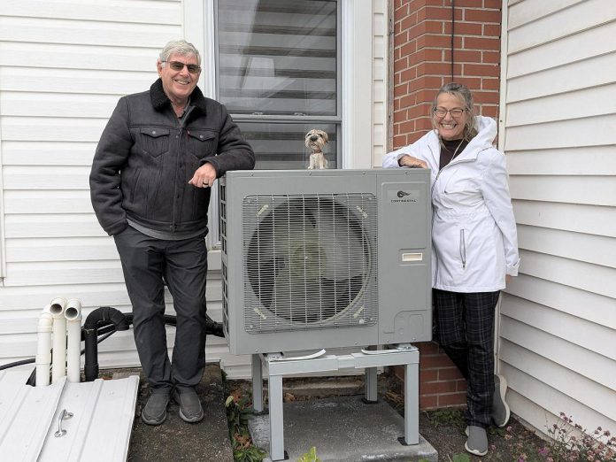 Rick and Annie Lochhead of Norwood stand next to the outdoor unit of their air source heat pump, which they installed in 2023. After doing some other upgrades, they estimate they're saving $50/month on their energy bills. (Photo: Clara Blakelock / GreenUP)