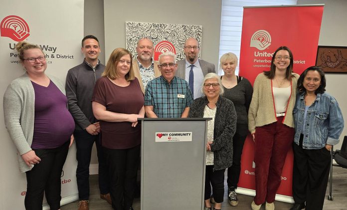 The United Way Peterborough & District's 2025 community campaign Paul Rellinger stands behind the podium with his wife Mary to his left, along with United Way CEO Jim Russell and board chair Marcus Harvey (both standing behind Rellinger) and United Way staff during an announcement at the United Way's office at 277 Stewart Street on May 21, 2025. (Photo: Jeannine Taylor / kawarthaNOW)