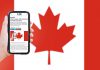 A hand holding a mobile phone showing kawarthaNOW's Canada Day hours story, with a Canadian flag in the background. (Photo: kawarthaNOW)