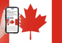 A hand holding a mobile phone showing kawarthaNOW's Canada Day hours story, with a Canadian flag in the background. (Photo: kawarthaNOW)