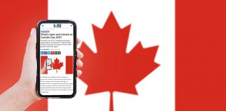 A hand holding a mobile phone showing kawarthaNOW's Canada Day hours story, with a Canadian flag in the background. (Photo: kawarthaNOW)