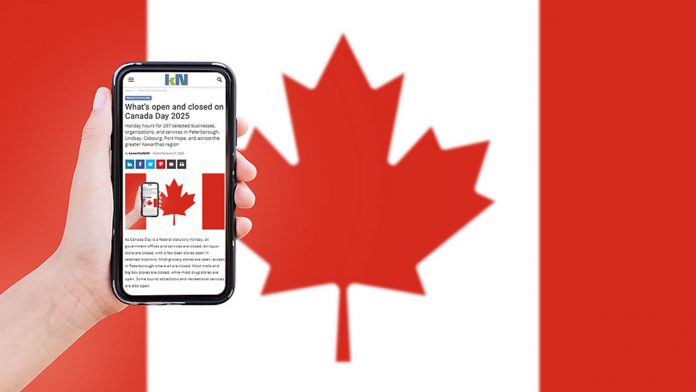 A hand holding a mobile phone showing kawarthaNOW's Canada Day hours story, with a Canadian flag in the background. (Photo: kawarthaNOW)