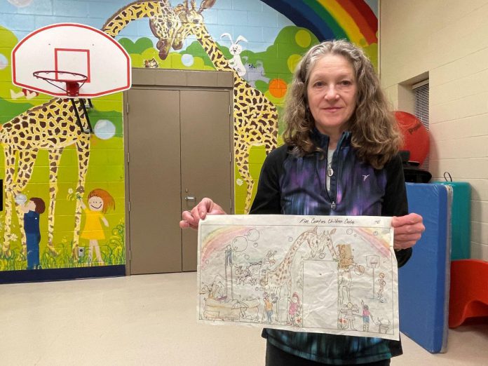Artist Sharon Pearsall displays the concept drawing she used to map out the giant mural that she created as a high school student on the gym wall at Five Counties Children's Centre in Peterborough in 1981. The mix of rainbows, giraffes, cats, children, bubbles and fun that are infused into the actual artwork are evident in the design. (Photo: Five Counties Children's Centre)