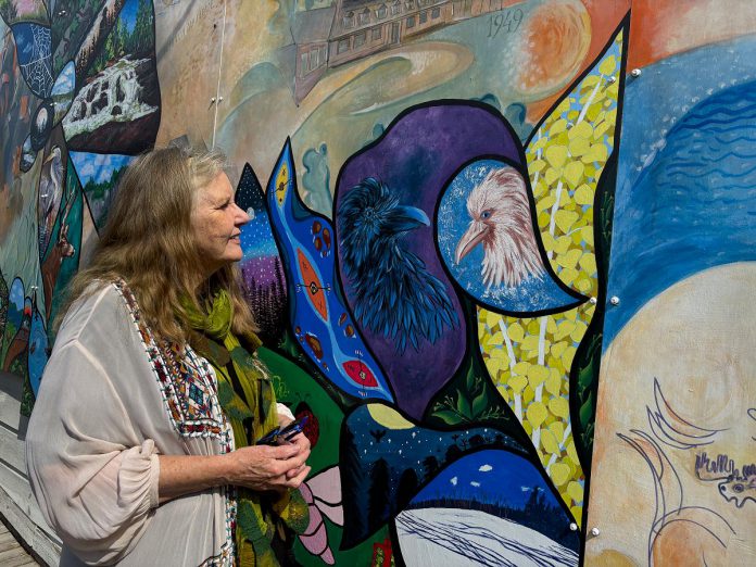 Artist and marketing director for A Place for the Arts in Bancroft, Gayle Crosmaz stands with the yin-yang-inspired white and black ravens she painted for the "Embrace" mural revitalization project at 23 Bridge Street. Crosmaz also designed the layout of the two large dragonflies and firefly that make up the foreground which were divided into smaller vignettes for community artists to paint images of what they love about Bancroft. (Photo: Megan Gallant / kawarthaNOW)