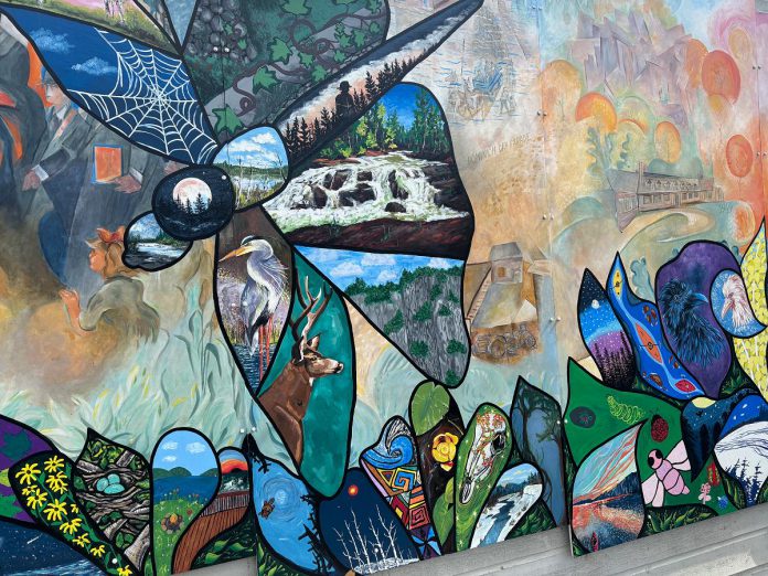 To revitalize the 2014 Arne Roosman mural at 23 Bridge Street in Bancroft, A Place for the Arts took inspiration from stain-glass designs by overlaying two large dragonflies, a firefly, and blades of glass with a black, thick outline. The new images were then divided into smaller sections that community members painted with images they liked about Bancroft. (Photo: Megan Gallant / kawarthaNOW)