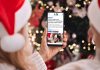 A person wearing a Santa hat in front of a Christmas tree holding a smartphone displaying the 2025 holiday hours listing on kawarthaNOW's website. (Photo: kawarthaNOW)