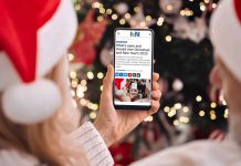 A person wearing a Santa hat in front of a Christmas tree holding a smartphone displaying the 2025 holiday hours listing on kawarthaNOW's website. (Photo: kawarthaNOW)
