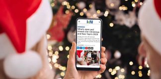 A person wearing a Santa hat in front of a Christmas tree holding a smartphone displaying the 2025 holiday hours listing on kawarthaNOW's website. (Photo: kawarthaNOW)