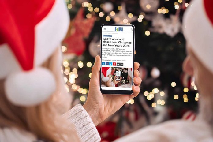 A person wearing a Santa hat in front of a Christmas tree holding a smartphone displaying the 2025 holiday hours listing on kawarthaNOW's website. (Photo: kawarthaNOW)