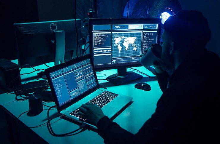 A hacker with a laptop and a secondary monitor showing data. (Stock photo)