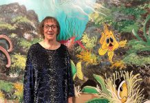 Artist Pat Calvert poses in front of a section of her mural at Five Counties Children's Centre in Peterborough, with Silly the Goldfish in the frame. Silly is a character that shows up around 500 times in various different situations and poses on the expansive, water-themed mural that has graced the walls and hallways of Five Counties for nearly 25 years. (Photo: Five Counties Children's Centre)