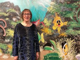 Artist Pat Calvert poses in front of a section of her mural at Five Counties Children's Centre in Peterborough, with Silly the Goldfish in the frame. Silly is a character that shows up around 500 times in various different situations and poses on the expansive, water-themed mural that has graced the walls and hallways of Five Counties for nearly 25 years. (Photo: Five Counties Children's Centre)