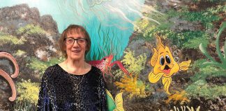 Artist Pat Calvert poses in front of a section of her mural at Five Counties Children's Centre in Peterborough, with Silly the Goldfish in the frame. Silly is a character that shows up around 500 times in various different situations and poses on the expansive, water-themed mural that has graced the walls and hallways of Five Counties for nearly 25 years. (Photo: Five Counties Children's Centre)