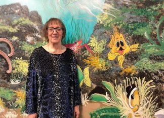 Artist Pat Calvert poses in front of a section of her mural at Five Counties Children's Centre in Peterborough, with Silly the Goldfish in the frame. Silly is a character that shows up around 500 times in various different situations and poses on the expansive, water-themed mural that has graced the walls and hallways of Five Counties for nearly 25 years. (Photo: Five Counties Children's Centre)