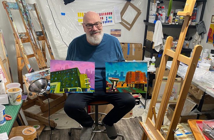 Artist David Geene in his home studio with two 12 by 16 inch paintings of existing buildings that will be featured in his "Economies of Scale?" solo exhibit at Watson & Lou in downtown Peterborough from April 4 to 25, 2026. Titled (left to right) "196 Hunter Street East" and "212 Brock Street from Hunter W. and Aylmer Streets," the works are two of the 12 acrylic paintings that aim to explore the politics and ethics of two proposed high-rise structures in Peterborough, at 90 Hunter Street and on Crescent Street. Including interpretations of the proposed buildings, the exhibit will launch with an opening reception from 2 to 4 p.m. on Saturday, April 4. (Photo courtesy of David Geene)