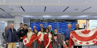 Rashid Sheikh Hassan (fourth from left, with Canadian flag) hugging his mother with his two sisters (wrapped in Canadian flags), brother (third from right, squatting), and father (far right) at Toronto's Pearson International Airport on April 24, 2026, pictured with members of the Azadi Peterborough group that sponsored the Hassan family to come to Canada and join their son in Peterborough. (Photo courtesy of Dave McNab)