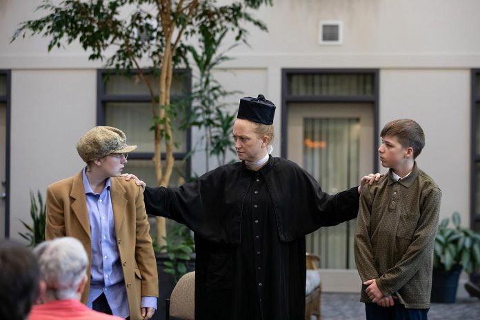 Sarah Myles (centre), a newcomer to the stage, performs the role of Father Leo, the parish priest of the altar boys Pete (June Covier) and Jimmy Clarke (Charlie Harris) during a special preview dress rehearsal of Trent Valley Archives Theatre's "The Kid from Simcoe Street" at Canterbury Gardens in Peterborough on May 28, 2026. In this scene, Father Leo reprimands the two boys after catching them in the middle of a prank. (Photo: Jordan Lyall Photography)