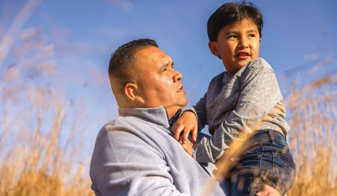 An Indigenous man holding a boy in his arms with a field in the background. (Photo: Alto)