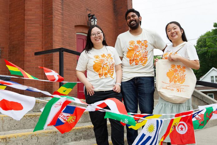 At an announcement on June 10, 2026 of the fifth annual Canadian Multicultural Festival presented by the New Canadians Centre, Peterborough Immigration Partnership Officer Michelle Nguyen, Manager of Wellness and Community Education Bhisham Ramoutar, and Fund Development Officer Asuka Ballantyne show off the branded merchandise that will be available for sale during the festival. Proceeds will help fund the non-profit organization's work to support immigrants, refugees, and other newcomers to the Kawarthas region. (Photo: Jordan Lyall / kawarthaNOW)