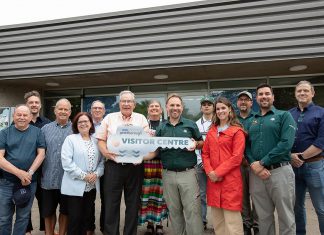 Peterborough mayor Jeff Leal (third from left, front) accepts a symbolic key from Parks Canada's manager of canal operations Chad Buchner for the new Peterborough Visitor Centre during a media event on June 25, 2026, the day before the centre opened to the public, surrounded by city councillors, Parks Canada staff, and two representatives of the Indigenous community. Operated by Peterborough Tourism, the visitor centre is located in the building previously operated by Parks Canada as the Peterborough Lift Lock Visitor Centre until 2020. (Photo: Jordan Lyall / kawarthaNOW)