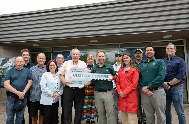 Peterborough mayor Jeff Leal (third from left, front) accepts a symbolic key from Parks Canada's manager of canal operations Chad Buchner for the new Peterborough Visitor Centre during a media event on June 25, 2026, the day before the centre opened to the public, surrounded by city councillors, Parks Canada staff, and two representatives of the Indigenous community. Operated by Peterborough Tourism, the visitor centre is located in the building previously operated by Parks Canada as the Peterborough Lift Lock Visitor Centre until 2020. (Photo: Jordan Lyall / kawarthaNOW)