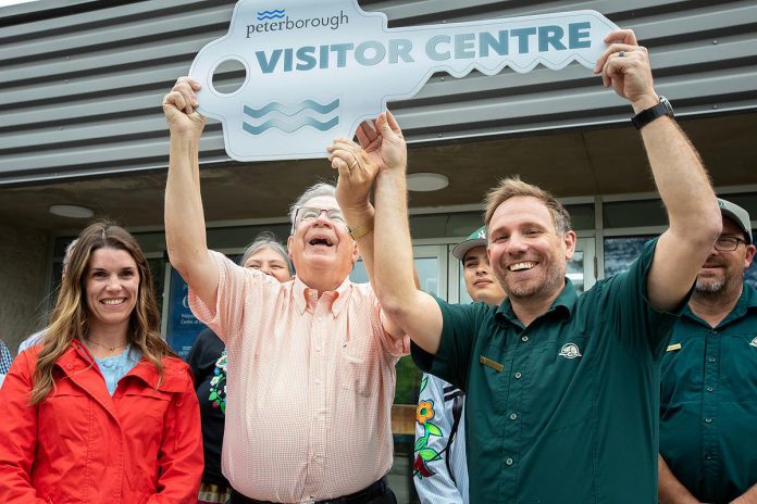 Peterborough mayor Jeff Leal and Parks Canada's manager of canal operations Chad Buchner lift a symbolic key for the new Peterborough Visitor Centre during a media event on June 25, 2026, with Peterborough MP Emma Harrison pictured at left. The city has entered into an agreement with Parks Canada to use the building that previously was the home of the Peterborough Lift Lock Visitor Centre until 2020. (Photo: Jordan Lyall / kawarthaNOW)