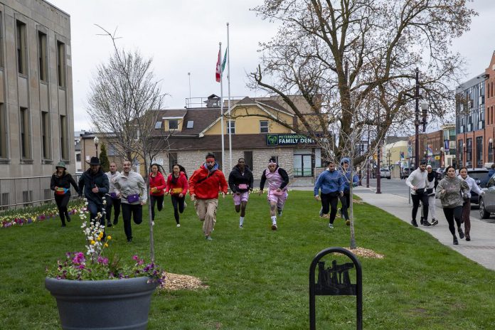 The Amazing Race Canada contestants race in front of Peterborough City Hall in downtown Peterborough in the fifth episode of the 12th season of the reality TV series, which was filmed in Peterborough and the Kawarthas in April and May 2026. Throughout the episode, teams of two will be competing in tasks at destinations across the region, including at The Canadian Canoe Museum, the Ontario Turtle Conservation Centre, Kawartha Dairy, and more. (Photo: Bell Media)