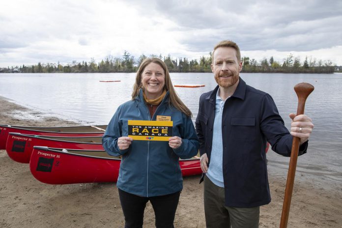 Carolyn Hyslop, executive director of The Canadian Canoe Museum in Peterborough, holds a clue for participants of The Amazing Race Canada with host Jon Montgomery at the beach at Beavermead Park in Peterborough. Airing on August 11, 2026 on CTV and streaming on Crave the next day, the fifth episode of the 12th season will include destinations across Peterborough and the Kawarthas, including the museum, the Ontario Turtle Conservation Centre in Peterborough County, Kawartha Dairy in Bobcaygeon, and Lock 31 of the Trent-Severn Waterway in Buckhorn. (Photo: Bell Media)