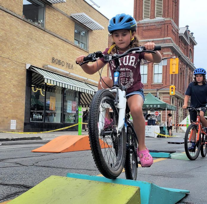 Parents shopped while kids rode on GreenUP's bike playground as the Second Saturday Sidewalk Sale turned a block of Charlotte Street in downtown Peterborough into a car-free zone on August 8, 2026. Canadian cities are dabbling with offering more space to pedestrians to create safer, less polluted, and more welcoming streets. (Photo: Brett Throop / GreenUP)