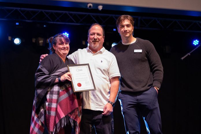 The Venue owners April and Kevin Sendel with a certificate of recognition of five years of family ownership presented by Peterborough and Kawarthas Chamber of Commerce member engagement coordinator Liam O'Connor during a celebration at the downtown Peterborough event space on October 13, 2026. (Photo: Joshua Gillis)